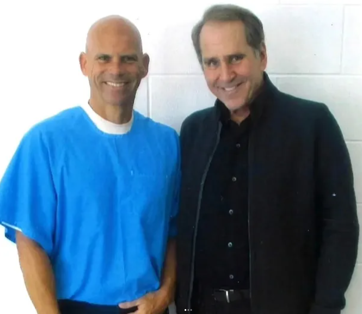 Robert Rand standing beside Lyle Menendez, who wears a blue prison uniform, both smiling against a white cinder-block wall.